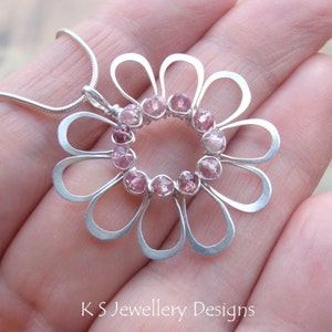 May include: A silver flower pendant necklace with pink gemstones set in the center. The pendant has a delicate, open design with petal-shaped loops. The necklace is on a silver chain. The text "KS Jewellery Designs" is at the bottom.