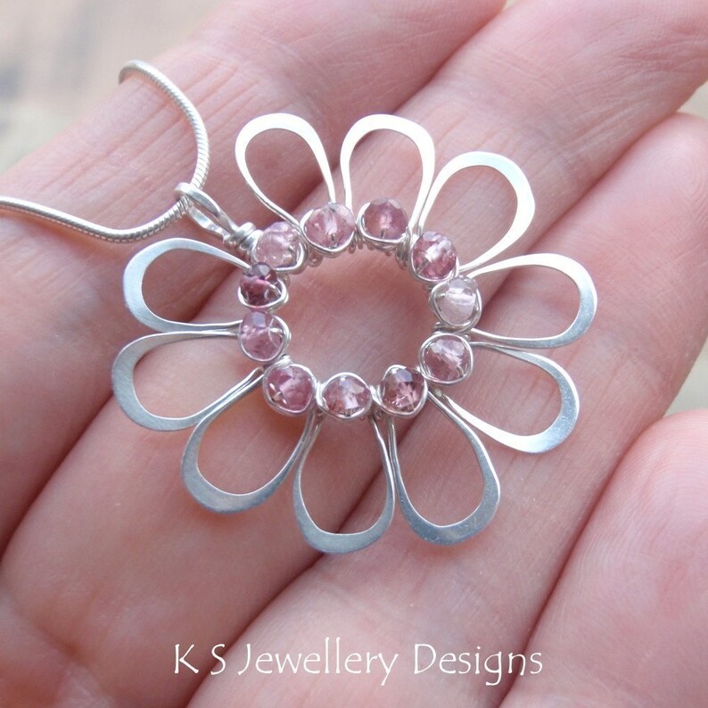 May include: A silver flower pendant necklace with pink gemstones set in the center. The pendant has a delicate, open design with petal-shaped loops. The necklace is on a silver chain. The text "KS Jewellery Designs" is at the bottom.