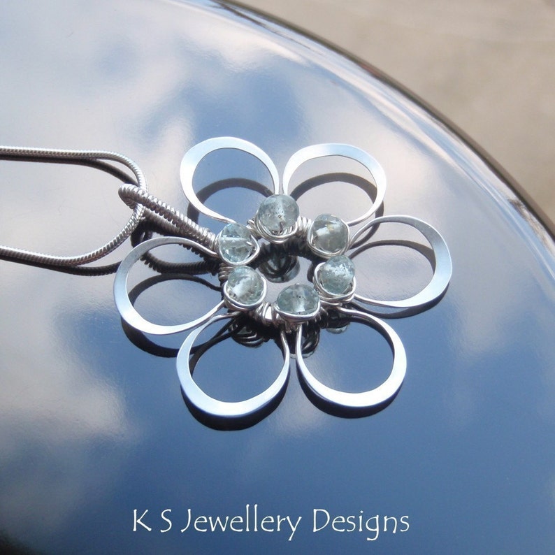 May include: A silver flower pendant necklace with six light blue gemstones at the center. The pendant has a floral design with six rounded petals. The necklace chain is a thin, silver snake chain. The text "K S Jewellery Designs" is at the bottom.