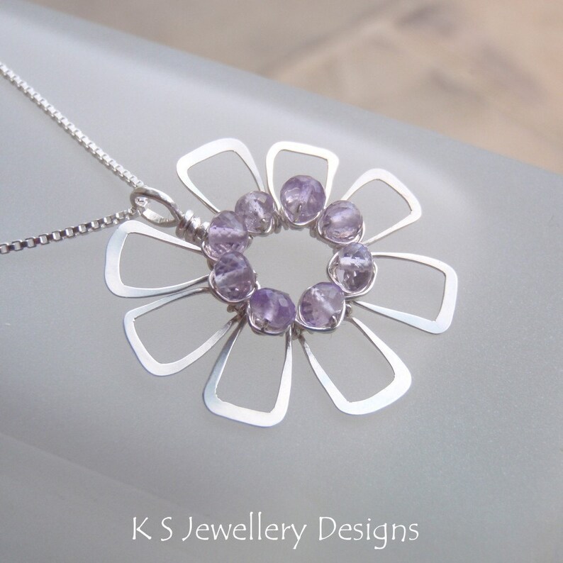 May include: A silver pendant necklace featuring a flower design. The pendant has a floral shape with open petals and a circle of small, light purple gemstones at the center. The necklace has a delicate chain. The text "K S Jewellery Designs" is at the bottom.
