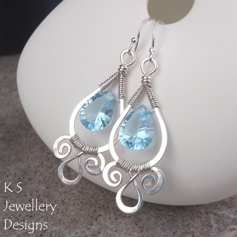 May include: Silver teardrop earrings with light blue teardrop-shaped crystals. The earrings have a delicate, intricate design with swirls.