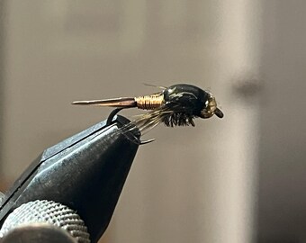 May include: A close-up of a fly fishing lure. The lure features a black body, gold wire wrapping, and brown feather details. It is held in a black vise, showcasing the intricate craftsmanship of the fly.