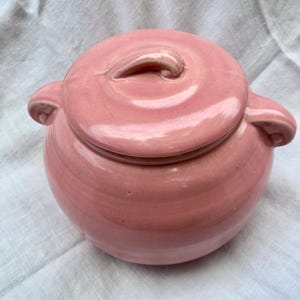 May include: A pink ceramic pot with a lid and two handles. The pot has a rounded shape and a smooth, glossy finish.