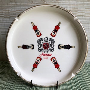 May include: White ceramic ashtray with a gold rim. The center features a crest and six illustrations of red and black bottles. The word "Arrow Cordials" is printed in red script.