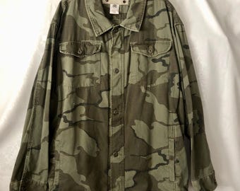 May include: A camouflage-patterned jacket in shades of green and brown. The jacket has a button-up front, two chest pockets with button closures, and two side pockets. The collar is a classic style, and the sleeves are long.