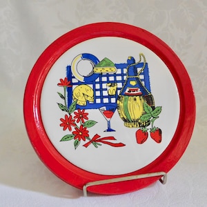 May include: A red and white ceramic coaster with a design of a wine bottle, grapes, cheese, and flowers. The coaster has a red rim and a white center.