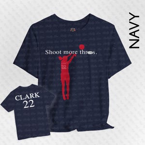 Puede incluir: Camisetas azul marino con temática de baloncesto. Una camiseta presenta una silueta roja de un jugador de baloncesto lanzando un balón, con el texto "Shoot more threes". La otra camiseta tiene "CLARK 22" impreso en la espalda.