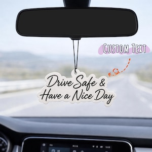 May include: A white, cloud-shaped car air freshener hanging from a rearview mirror by a black string. The air freshener features black cursive text that reads Drive Safe & Have a Nice Day. A small graphic indicates a space for custom text.