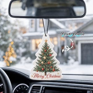 May include: A white, tree-shaped car air freshener hanging from a rearview mirror. The ornament features a printed illustration of a decorated Christmas tree with red and gold ornaments, a star topper, and the red text Merry Christmas at the bottom.