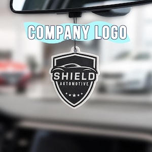 May include: A black and white shield-shaped car air freshener hanging from a rearview mirror. The design features a stylized car silhouette above the text SHIELD AUTOMOTIVE and five small stars at the bottom, all printed on a textured surface.