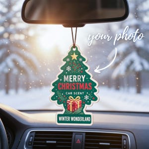 May include: A Christmas tree-shaped car air freshener hanging from a rearview mirror. The green ornament features a gold star, red gift box, and white text reading MERRY CHRISTMAS CAR SCENT and WINTER WONDERLAND, with a white arrow pointing to your photo.