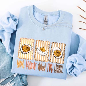 May include: A light blue sweatshirt with a graphic of three deviled eggs on striped backgrounds. The text "You know why I'm here" is printed below the eggs in orange and brown. A silver necklace with a flower pendant is also visible.