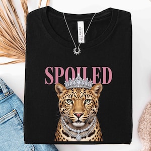 May include: Black t-shirt with the word "SPOILED" in pink letters above a leopard wearing a crown and a pearl necklace. A silver necklace with a sunburst pendant is also visible.