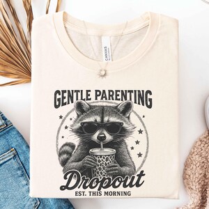 May include: Cream-colored t-shirt featuring a raccoon wearing sunglasses and drinking from a leopard-print cup. The text reads "GENTLE PARENTING Dropout EST. THIS MORNING" in a circular design. The shirt is folded.