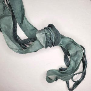 Puede incluir: Una larga cinta fluida en tonos verde azulado y gris, anudada en el centro. Los bordes están acabados con un delicado detalle cosido. La textura de la cinta parece suave y ligeramente translúcida, con un brillo sutil.