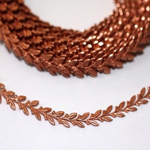 May include: A roll of copper-colored leaf trim, with a detailed, repeating pattern. The trim is in focus, with a portion coiled in the background. The leaves are small and delicate, creating a decorative element. This trim is suitable for various craft projects.