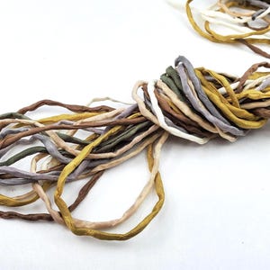Könnte beinhalten: Ein Bündel aus gedrehtem Seidenband in verschiedenen Erdtönen, darunter Braun, Olivgrün, Gold und Grau. Das Band ist eng gewickelt und scheint handgefertigt zu sein, mit einer leicht strukturierten Oberfläche. Ideal für Bastelarbeiten und Verzierungen.