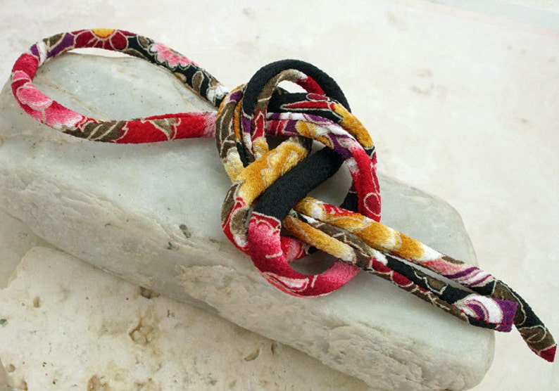 May include: A colorful fabric cord with a floral pattern in shades of red, pink, black, brown, and gold. The cord is tied in a knot and is lying on a white surface.