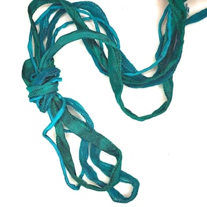 May include: A collection of intertwined fabric ribbons in various shades of teal, turquoise, and deep emerald green. The ribbons feature a mix of smooth, satin-like textures and frayed, woven edges, gathered together in a loose, knotted arrangement.