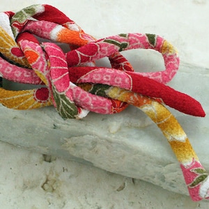 Könnte beinhalten: Ein farbenfrohes Stoffband, das zu einem Knoten gebunden ist und ein rotes, rosa, gelbes und grünes Blumenmuster aufweist. Das Band liegt auf einer hellgrauen Steinoberfläche.