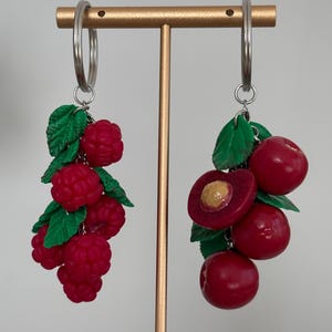 May include: A pair of silver hoop earrings with dangling clusters of red raspberries and cherries. The earrings feature detailed, realistic fruit designs with green leaves, showcasing a playful, nature-inspired theme.