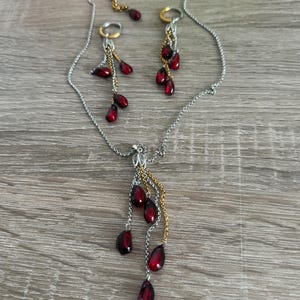 K&ouml;nnte beinhalten: Ein silberfarbenes Halsketten- und Ohrring-Set. Die Halskette und die Ohrringe weisen eine Kaskade aus dunkelroten, tropfenf&ouml;rmigen Perlen auf. Die Ohrringe haben goldfarbene Akzente, und die Halskette hat einen dekorativen Anh&auml;nger mit goldfarbenen Details.