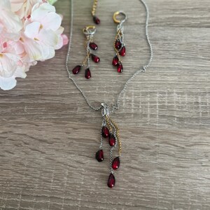 K&ouml;nnte beinhalten: Ein silberfarbenes Halsketten- und Ohrring-Set. Der Schmuck hat tropfenf&ouml;rmige, dunkelrote Perlen. Die Halskette und die Ohrringe haben goldfarbene Kettenakzente. Der Schmuck wird auf einer Holzoberfl&auml;che pr&auml;sentiert.