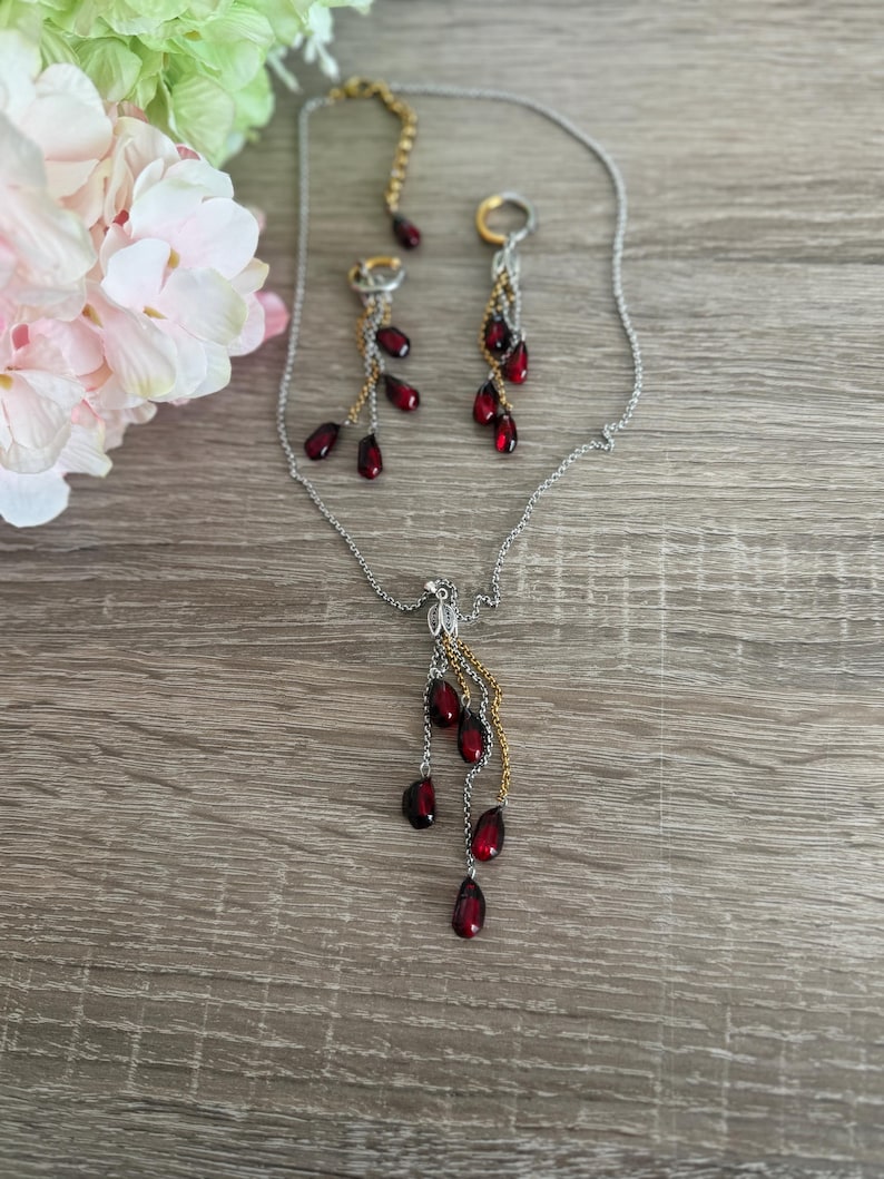 K&ouml;nnte beinhalten: Ein silberfarbenes Halsketten- und Ohrring-Set. Der Schmuck hat tropfenf&ouml;rmige, dunkelrote Perlen. Die Halskette und die Ohrringe haben goldfarbene Kettenakzente. Der Schmuck wird auf einer Holzoberfl&auml;che pr&auml;sentiert.
