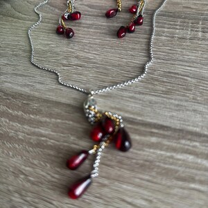 K&ouml;nnte beinhalten: Ein silberfarbenes Halsketten- und Ohrring-Set. Die Halskette hat einen Anh&auml;nger mit roten, tropfenf&ouml;rmigen Perlen. Die Ohrringe haben ebenfalls rote Perlen. Der Schmuck wird auf einer Holzoberfl&auml;che pr&auml;sentiert.