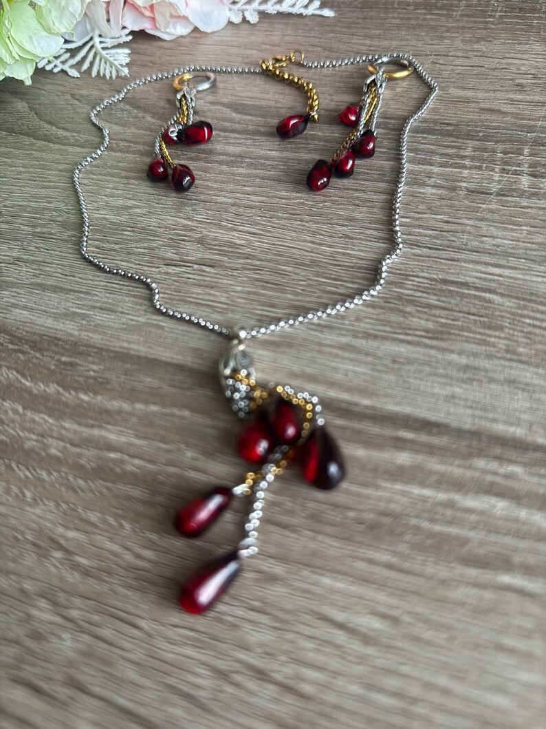 K&ouml;nnte beinhalten: Ein silberfarbenes Halsketten- und Ohrring-Set. Die Halskette hat einen Anh&auml;nger mit roten, tropfenf&ouml;rmigen Perlen. Die Ohrringe haben ebenfalls rote Perlen. Der Schmuck wird auf einer Holzoberfl&auml;che pr&auml;sentiert.