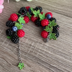 May include: A handcrafted bracelet with a design of red raspberries, black blackberries, and blue blueberries. The bracelet includes green leaf accents and a silver chain with a leaf charm. The berries are clustered together to form the bracelet.