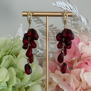 Könnte beinhalten: Ein Paar goldfarbene Ohrringe mit Anhängern aus dunkelroten, granatapfelförmigen Perlen. Die Ohrringe haben ein dekoratives Filigranmuster an der Oberseite und werden vor einem Blumenvorhang präsentiert.