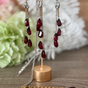 K&ouml;nnte beinhalten: Ein Schmuckset mit Halskette und Ohrringen zum Thema Granatapfel. Die Ohrringe und die Halskette haben silberne Ketten mit herabh&auml;ngenden, dunkelroten, tropfenf&ouml;rmigen Perlen. Der Schmuck wird auf einem goldfarbenen St&auml;nder pr&auml;sentiert.