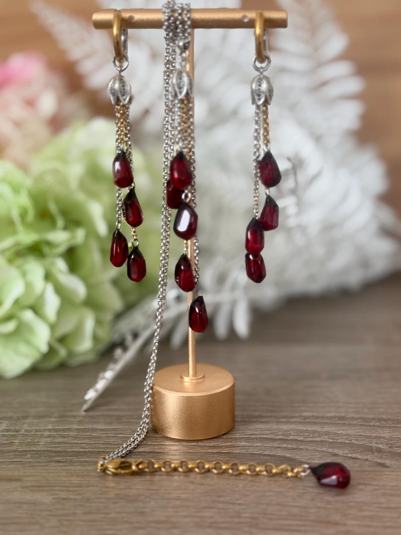 K&ouml;nnte beinhalten: Ein Schmuckset mit Halskette und Ohrringen zum Thema Granatapfel. Die Ohrringe und die Halskette haben silberne Ketten mit herabh&auml;ngenden, dunkelroten, tropfenf&ouml;rmigen Perlen. Der Schmuck wird auf einem goldfarbenen St&auml;nder pr&auml;sentiert.