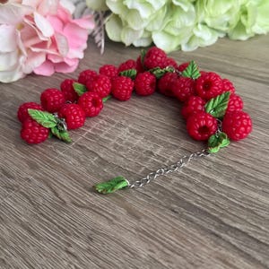 Könnte beinhalten: Ein Armband mit leuchtend roten Himbeerperlen und grünen Blättern, verbunden durch eine silberne Kette. Die Himbeeren sind gruppiert, wodurch ein lebendiges Accessoire entsteht. Das Armband ist ein einzigartiges Schmuckstück.