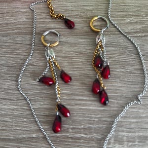 K&ouml;nnte beinhalten: Eine silberfarbene Halskette und ein passendes Ohrring-Set. Die Ohrringe haben gold- und silberfarbene Reifen mit herabh&auml;ngenden roten tropfenf&ouml;rmigen Perlen. Die Halskette hat eine silberne Kette mit einem goldfarbenen Verschluss und einem einzelnen roten Perlenanh&auml;nger.