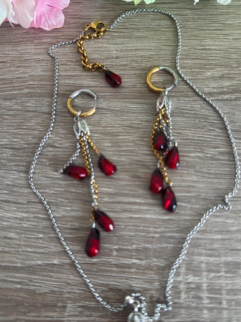 K&ouml;nnte beinhalten: Eine silberfarbene Halskette und ein passendes Ohrring-Set. Die Ohrringe haben gold- und silberfarbene Reifen mit herabh&auml;ngenden roten tropfenf&ouml;rmigen Perlen. Die Halskette hat eine silberne Kette mit einem goldfarbenen Verschluss und einem einzelnen roten Perlenanh&auml;nger.