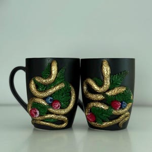 May include: Two matte black ceramic mugs featuring three-dimensional gold snakes coiled around green fern leaves and small red and blue berries. The snakes have a textured scale pattern, and the mugs have a smooth, dark finish with a handle on the left.
