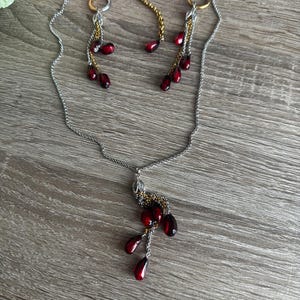 K&ouml;nnte beinhalten: Ein silberfarbenes Halsketten- und Ohrring-Set mit passenden Ohrringen. Der Schmuck hat dunkelrote, tropfenf&ouml;rmige Perlen. Die Ohrringe und die Halskette haben goldfarbene Akzente und liegen auf einer Holzoberfl&auml;che.