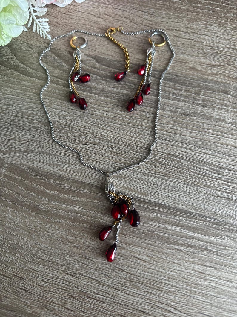 K&ouml;nnte beinhalten: Ein silberfarbenes Halsketten- und Ohrring-Set mit passenden Ohrringen. Der Schmuck hat dunkelrote, tropfenf&ouml;rmige Perlen. Die Ohrringe und die Halskette haben goldfarbene Akzente und liegen auf einer Holzoberfl&auml;che.