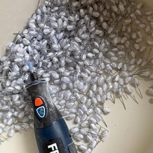 K&ouml;nnte beinhalten: Nahaufnahme einer Ansammlung kleiner, klarer, tropfenf&ouml;rmiger Perlen mit silbernen Drahtstielen. Ein blaues und schwarzes Rotationswerkzeug mit der Marke "FERM" befindet sich im Vordergrund. Die Perlen befinden sich in einem hellen Beh&auml;lter.