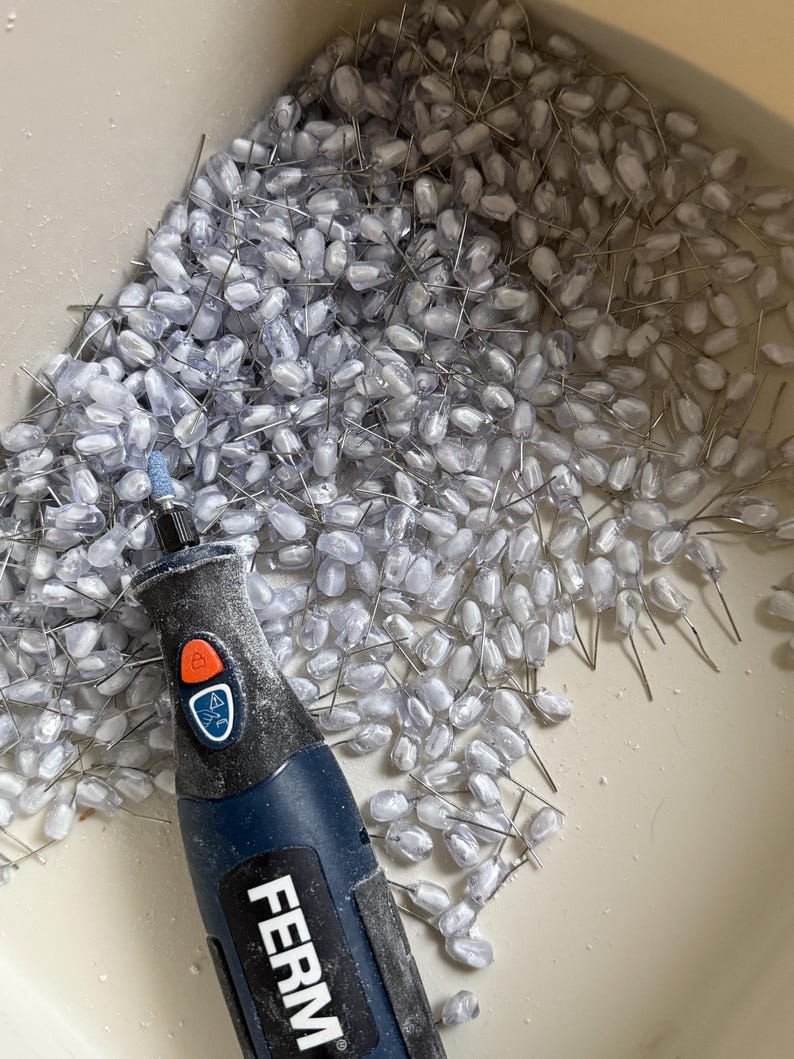 K&ouml;nnte beinhalten: Nahaufnahme einer Ansammlung kleiner, klarer, tropfenf&ouml;rmiger Perlen mit silbernen Drahtstielen. Ein blaues und schwarzes Rotationswerkzeug mit der Marke "FERM" befindet sich im Vordergrund. Die Perlen befinden sich in einem hellen Beh&auml;lter.