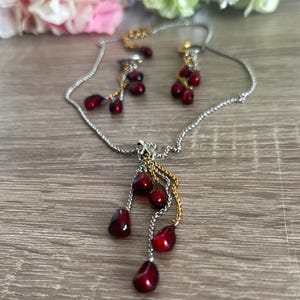 K&ouml;nnte beinhalten: Ein silberfarbenes Halsketten- und Ohrring-Set mit Granatapfel-Design. Die Halskette und die Ohrringe haben h&auml;ngende, dunkelrote, tropfenf&ouml;rmige Perlen. Die Halskette hat eine silberne Kette mit goldenen Akzenten. Die Ohrringe haben ein &auml;hnliches Design. Das Schmuckst&uuml;ck wird auf einer Holzoberfl&auml;che pr&auml;sentiert.