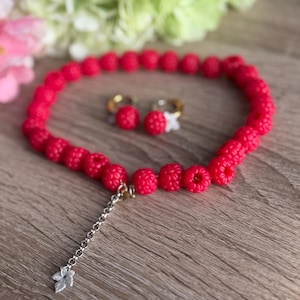 May include: A bright red raspberry jewellery set, comprising a necklace and matching earrings. The necklace has a chain with a leaf charm. The earrings feature gold hoops. The jewellery is set against a wooden surface.