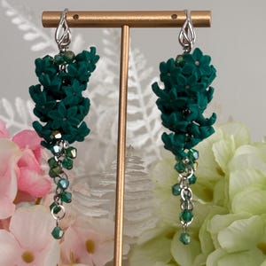 May include: A pair of dangling earrings with a floral design. The earrings feature dark green flower clusters and dangling chains with green beads. The ear wires are silver-colored. The earrings are displayed on a gold-colored stand.