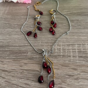 K&ouml;nnte beinhalten: Ein silbernes Halsketten- und Ohrring-Set mit goldenen Akzenten. Der Schmuck hat tropfenf&ouml;rmige, dunkelrote Perlen. Die Halskette und die Ohrringe werden auf einer Holzoberfl&auml;che mit rosa und gr&uuml;nen Blumen im Hintergrund pr&auml;sentiert.