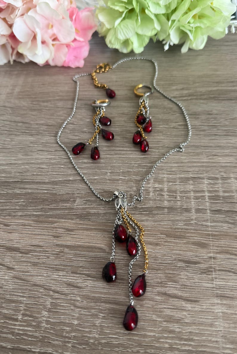K&ouml;nnte beinhalten: Ein silbernes Halsketten- und Ohrring-Set mit goldenen Akzenten. Der Schmuck hat tropfenf&ouml;rmige, dunkelrote Perlen. Die Halskette und die Ohrringe werden auf einer Holzoberfl&auml;che mit rosa und gr&uuml;nen Blumen im Hintergrund pr&auml;sentiert.