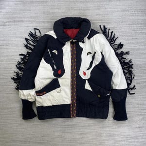 Peut inclure: Veste noire et blanche ornée de têtes de chevaux. La veste a un col, des poignets et des franges noirs. La fermeture éclair est ornée d'une bordure à motifs. La doublure intérieure est rouge.