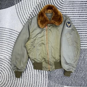 Könnte beinhalten: Olivgrüne Bomberjacke mit braunem Kunstfellkragen. Sie hat einen Reißverschluss vorne, gerippte Bündchen und Saum sowie einen Army Air Forces-Aufnäher auf dem Ärmel. Die Jacke wird auf einer gemusterten Oberfläche präsentiert.