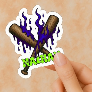 May include: A sticker featuring two crossed baseball bats with a wood grain texture, overlaid with dripping purple liquid. The word "NATHAN" is written in green, stylized lettering.